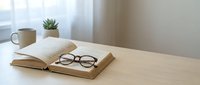 Open book with eyeglasses on wooden desk near window, featuring succulent plant and mug. Suitable for education, reading, and workspace design projects.