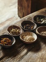 Assorted dried herbs and spices in rustic wooden bowls on weathered table surface, moody natural lighting creates atmospheric composition for culinary concepts.
