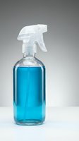 Clear glass spray bottle with blue cleaning liquid and white trigger sprayer on neutral background for household cleaning and disinfection purposes.