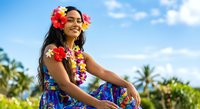 Young woman of South Asian descent wearing vibrant floral lei and tropical dress, smiling outdoors with palm trees and blue sky, perfect for travel, vacation, and summer lifestyle content.
