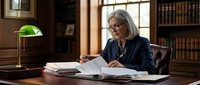 Senior businesswoman with gray hair reviewing documents at wooden desk in traditional office library with banker lamp and bookshelves background.