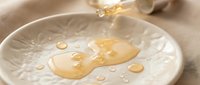 Honey dripping onto white ceramic plate with water droplets, close-up of golden liquid pouring in studio setting.