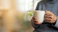 Person of African descent wearing dark gray turtleneck sweater holding white ceramic mug, warm indoor lighting with blurred background, suitable for beverage, lifestyle and wellness marketing.