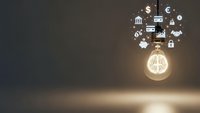 Glowing lightbulb with financial icons floating above on dark background representing business innovation, investment ideas and economic growth concepts.