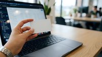 Female hand holding blank white business card near laptop with open software interface at wooden desk in modern office environment with blurred colleagues in background.