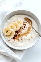 Greek yogurt bowl with fresh banana slices, peanut butter swirl and cocoa powder sprinkled on top, served in white ceramic bowl on marble surface.