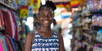 Young African American woman with bright smile wearing floral pattern dress standing in retail store. Colorful shopping environment with clothing racks and merchandise in background.