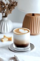 Latte art in ribbed glass on metal coaster against white background with minimal decor elements. Cozy coffee moment with gradient foam and leaf pattern.