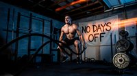 Shirtless male athlete with muscular build performing battle rope exercise in industrial gym setting with motivational signage and weight plates visible.