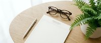 Blank notepad with pen and tortoiseshell glasses on a round wooden table beside a green fern plant, minimal home office workspace setup.