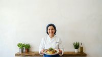 Chef in white uniform holding plate of fresh pasta salad with vegetables in modern kitchen studio setting.