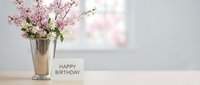 Pink cherry blossoms in silver vase beside happy birthday card on wooden table, soft bright background for spring celebration greeting.