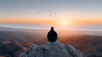 Person sitting on mountain peak watching sunrise over layered hills with flying birds in peaceful morning light for meditation and wellness content.