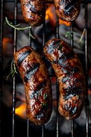 Grilled sausages with caramelized glaze and fresh herbs cooking on barbecue grill grates with visible flames, perfect for food marketing, menus, and cooking blogs.