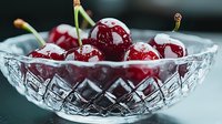 Fresh ripe cherries with water drops in crystal glass bowl on dark background, macro food photography showing texture and reflection.