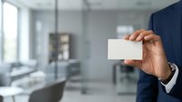 Businessman in dark suit holding blank white business card in modern office environment with glass partitions and natural light from windows.