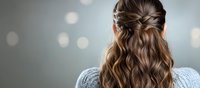 Young woman with elegant braided hairstyle wearing grey sweater against blurred bokeh background, view from behind, showing trendy half-up half-down style.