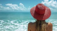 Woman wearing red sun hat gazing at turquoise ocean waves from beach. Back view of blonde female tourist enjoying tropical seascape under summer sky with fluffy clouds.
