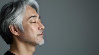 Side profile of mature Asian man with gray hair and beard against dark background, eyes closed in peaceful meditation or contemplation, expressing serenity and mindfulness.