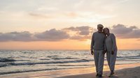 Senior couple walking together on beach at sunset enjoying romantic evening stroll by ocean waves with golden sky reflecting on water creating peaceful moment.