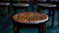 Vintage wooden bar stool with rich patina and distressed surface texture in dark moody lighting, showing natural wear patterns and aged character in rustic setting.