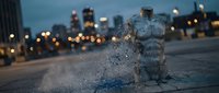 Splashing water from urban fountain at dusk with blurred city lights and skyscrapers in background, suitable for urban lifestyle and environmental design projects.