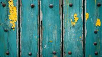 Weathered turquoise wooden door with peeling yellow paint and rustic metal rivets shows vintage texture and patina. Architectural detail creates rustic charm.