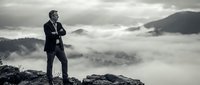 Professional businessman in suit standing on mountain peak above clouds contemplating success and achievement in dramatic landscape.