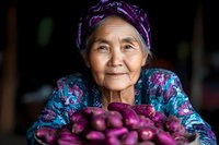 Senior Asian woman in traditional headscarf and floral clothing smiling while presenting fresh purple eggplants from harvest, authentic rural lifestyle portrait.