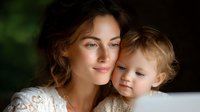 Tender moment between young Caucasian mother and toddler child with curly blonde hair, both smiling warmly in soft natural lighting against dark background.