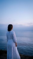 Young woman in white dress standing by ocean at sunset contemplating peaceful seascape horizon for wellness and mindfulness concepts.