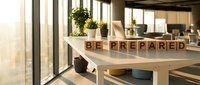 Wooden blocks spelling "BE PREPARED" on desk in modern office workspace with natural light, plants, and contemporary furniture for business motivation and planning concepts.