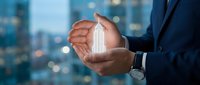 Businessman hands protecting glowing building hologram against blurred city skyline at dusk. Real estate investment and property protection concept.