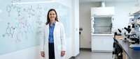 Professional female scientist in white lab coat standing confidently in modern research laboratory with molecular diagrams on whiteboard behind her.