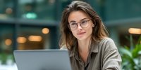 Young Caucasian businesswoman with glasses working on laptop in modern office environment, professional portrait for corporate marketing materials.