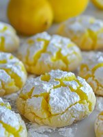 Crinkle lemon cookies dusted with powdered sugar and fresh citrus zest, showcasing distinctive cracked surface texture and bright yellow color. Fresh lemons in background.