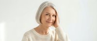 Mature woman with gray hair and fair complexion wearing cream knit sweater, hand near face, smiling at camera against white background for portrait and lifestyle use.