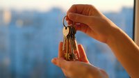 Hand holding house keys against blurred city skyline background at sunset, real estate property ownership concept for new homeowners and rental market.