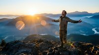 Young Caucasian male hiker stands arms outstretched on mountain summit at sunrise, fog-filled valley below, golden light flaring.