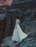Young woman in flowing white wedding dress standing on dramatic rocky cliff landscape at twilight for romantic bridal photography and fashion concepts.