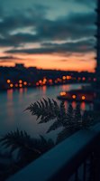Silhouette of fern leaves against blurred city lights and orange sunset sky reflected in calm water, creating moody atmospheric evening landscape with bokeh effect.
