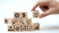 Hand placing wooden cube with lightbulb icon completing business strategy pyramid with financial and innovation symbols on white background.