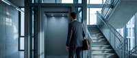 Professional businessman in suit walking towards modern elevator in contemporary office building with glass windows and metal staircase architecture.
