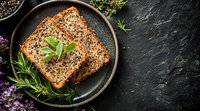 Homemade multigrain bread slices garnished with fresh herbs and seeds on black ceramic plate against dark stone background with copy space. Food photography.