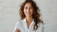 Young Mediterranean woman with curly brown hair and natural smile wearing white button shirt against light background. Professional headshot portrait.