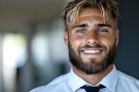 Confident young Caucasian businessman with beard and blonde hair wearing white shirt and tie smiling at camera in modern office environment. Professional portrait.