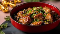 Glazed chicken wings in red ceramic bowl garnished with sesame seeds and green onions, served on dark surface with festive bokeh lights in background.