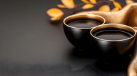 Two black ceramic cups of hot tea or coffee on dark background with golden autumn leaves and fabric, creating cozy atmosphere for relaxation.