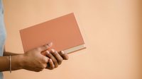 Person with medium skin tone holding closed orange book against neutral beige background, suitable for education, learning, reading, and publishing concepts.