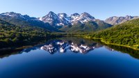 Majestic snow-capped mountain peaks reflecting in calm blue lake waters surrounded by lush green forest slopes, creating mirror-like symmetry in pristine wilderness.